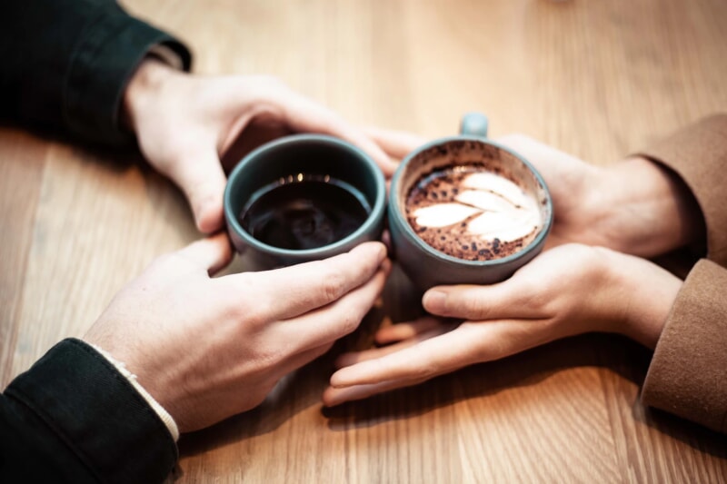 A pair of hands across from each other, each hold a cup of coffee.
